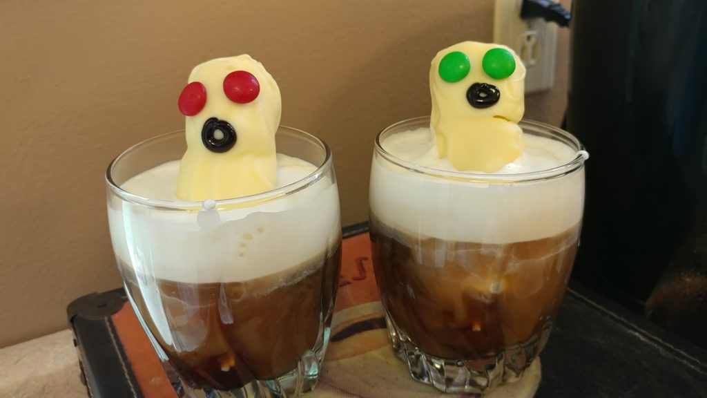 Two glass tumblers containing Irish coffee with whipped cream and white chocolate coated marshmallow ghouls with m&m eyes and a black mouth.